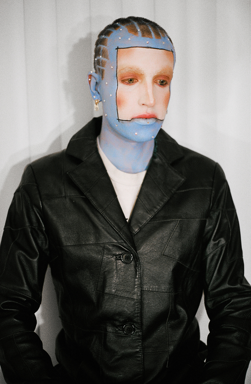 Waist-up photo of a model with blue skin and and a white square in the center of their face wearing a black leather jacket in front of a white background